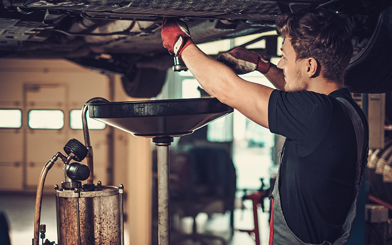 Professional technician performing under-car fluid service in an automotive repair shop.